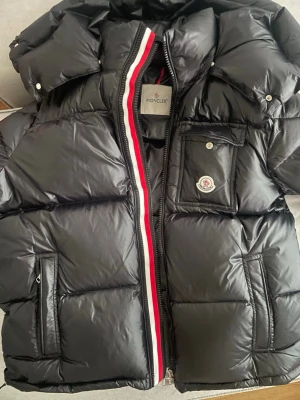 Svart Moncler pufferjacka strl 5 - Svart pufferjacka från Moncler med glansig finish och klassisk logga på bröstfickan. Jackan har dragkedja med röd och vit kant, flera fickor och hög krage. Perfekt för kalla dagar när du vill ha både stil och värme. Den kan också skannas både på Logan och tagen 