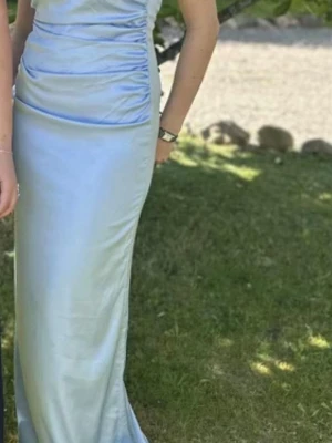 Ljusblå långklänning med öppen rygg - Säljer en elegant ljusblå långklänning med smala axelband och snygg drapering på framsidan. Klänningen har en öppen rygg och är tillverkad i ett glansigt, satinliknande material som ger en lyxig känsla. Perfekt för speciella tillfällen. Den är endast använd en gång och är i ett fint skick, pris kan diskuteras!
