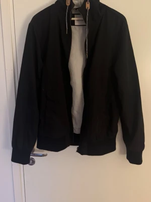 Svart vindjacka med dragkedja - Svart vindjacka från yourturn med hög krage och snörning. Jackan har dragkedja framtill och ribbade muddar vid ärmslut och nederkant. Perfekt för blåsiga dagar och enkel att matcha med olika outfits.