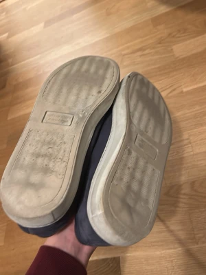 Svarta sneakers med vit sula - Säljer ett par svarta sneakers med vit platt sula och rund tå. Skorna har snörning och är tillverkade i tygmaterial Mocka,  med en enkel och stilren design. Insidan är gul och de passar perfekt för dig som gillar klassiska sneakers med låg profil.