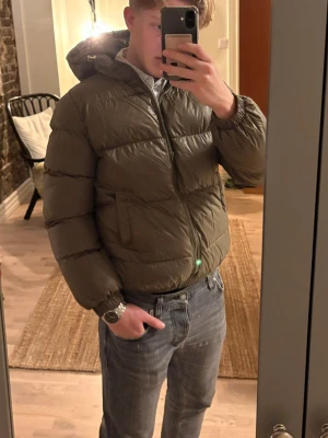 Olivgrön pufferjacka med huva - Säljer en sprillans ny olivgrön pufferjacka med huva och dragkedja framtill. Jackan har en klassisk pufferlook med breda kanaler och elastiska muddar vid ärmsluten. Perfekt för kyliga dagar och riktigt skön att bära. Storlek: M passar S