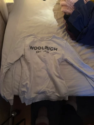 Vit sweatshirt från Woolrich - Vit långärmad sweatshirt från Woolrich med tryckt logga och text 'EST. 1830' på bröstet. Tröjan har rund hals och ribbade muddar vid ärmslut och nederkant. Perfekt för chill dagar och enkel att matcha med jeans eller mjukisbyxor.
