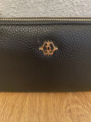 Svart plånbok med gulddetaljer från N - Snygg svart plånbok i skinn med guldfärgad logga och dubbla dragkedjor. Plånboken har en praktisk handledsrem och stilren design med struktur i materialet. Perfekt för att hålla koll på kort och kontanter när du är på språng.