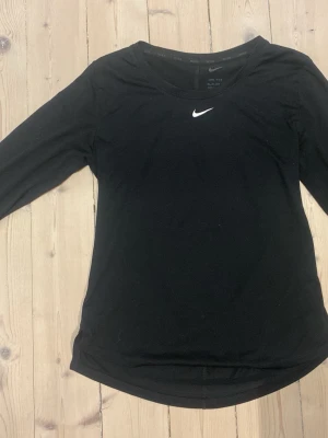 Svart långärmad Nike Dri-Fit träningströja - Svart långärmad träningströja från Nike. Tröjan har en rund hals och ett litet vitt Nike Swoosh-tryck på bröstet. Materialet är lätt och andas, perfekt för intensiva träningspass eller sportaktiviteter.