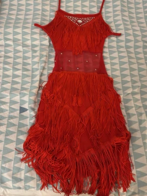 Röd fransklänning med strassdetaljer - Säljer en röd klänning med fransar och transparent midjeparti dekorerat med strass. Klänningen knyts i ryggen med snören och medföljande accessoarer: strasshalsvand, långa röda handskar med strass och matchande shorts. Perfekt för att sticka ut med coola detaljer och färg. Passar storlek 150