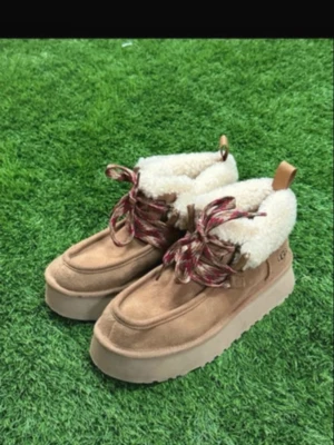 Beige boots från UGG med fuskpäls - Säljer ett par beige boots från UGG med fluffig fuskpäls på insidan och grov plattformssula. Skorna har snörning med röda och vita mönstrade skosnören och är tillverkade i mocka. Perfekta för kalla dagar och har en riktigt cozy vibe.