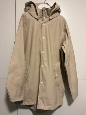 Beige regnjacka/regnkappa från Everest med huva - Snygg beige regnjacka/regnkappa från Everest med huva och vita knappar framtill. Jackan har två fickor, reflexdetaljer på ryggen och är tillverkad i syntetmaterial som står emot regn.