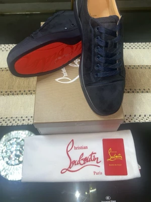Mörkblå mocka sneakers från Louboutin - Säljer ett par mörkblå sneakers från Christian Louboutin i lyxig mocka med klassisk röd sula. Skorna har rund tå, snörning och diskreta sömmar för en clean look. Perfekta för dig som vill ha något exklusivt och stilrent i garderoben.