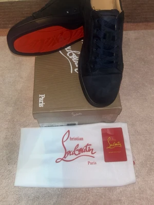 Christian Louboutin svarta sneakers - Svarta sneakers från Christian Louboutin med klassisk röd sula och rund tå. Skorna har snörning och är tillverkade i mocka med diskreta detaljer. Snygg och stilren design som sticker ut med den ikoniska röda undersidan. Levereras med originalkartong och dustbag.