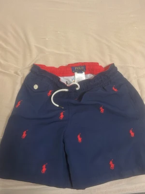 Blå badshorts från Polo Ralph Lauren - Snygga marinblå badshorts från Polo Ralph Lauren med röd broderad logga över hela tyget. Shortsen har röd resår i midjan, vit snörning och en ficka med knapp bak. Perfekta för stranden eller poolen!
