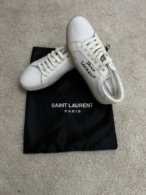 Saint Laurent sneakers - Säljer ett par ÄKTA vita sneakers från Saint Laurent med handskriven logga på sidan. Skorna har rund tå, platt sula och är tillverkade i canvas med vita snören. Klassisk och stilren design med diskreta detaljer. Storlek 41 sitter som 41. Skick 10/10. Endast dustbagen och själva skorna medföljer.