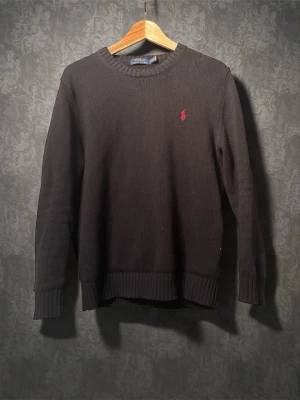 Svart stickad tröja från Polo Ralph Lauren - Svart stickad tröja från Polo Ralph Lauren med klassisk rund hals och röd broderad logga på bröstet. Tröjan har ribbade muddar vid ärmslut och nederkant samt långärmad passform. Perfekt för lager-på-lager under kyliga dagar.