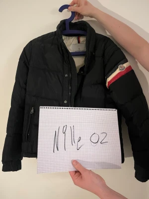 Svart pufferjacka från Moncler - Svart pufferjacka från Moncler med krage och dragkedja framtill. Jackan har en cool detalj med Moncler-logga och röd, vit och marinblå rand på ena ärmen. Insidan är ljus och har en innerficka med dragkedja. Perfekt för kalla dagar och riktigt snygg stil. Den är köpt från kaspersheat så kvitto finns och storleken är 1 (S)