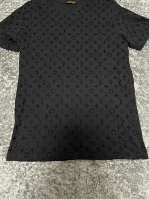 Svart Louis Vuitton T-shirt med monogram - Säljer en svart T-shirt från Louis Vuitton med det klassiska LV-monogrammet över hela plagget. T-shirten är tillverkad i 100% bomull och har en stilren, mörk look som passar dig som gillar exklusiva märken och snygg design.