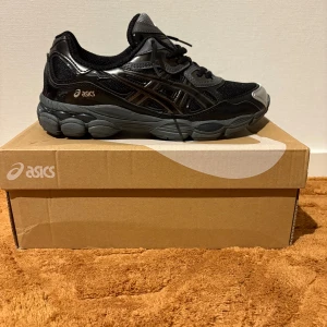 Svarta och grå ASICS sneakers strl 40 - ASICS sneakers i svart och grått med silvriga detaljer och coolt motiv på innersulan. Skorna har snörning, rund tå och platt sula. Materialmix av syntet och mesh ger både stil och komfort. Perfekt för dig som gillar sportig vibe och snygga detaljer.