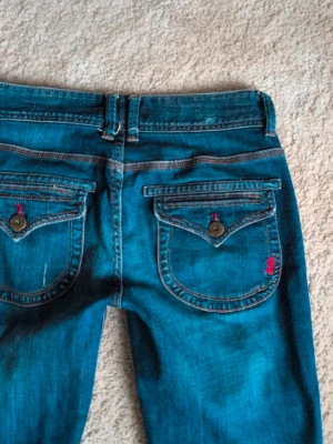 Blå True Boot jeans med ficklock - Säljer ett par blå jeans från True Boot med snygga ficklock och knappdetaljer bak. Jeansen har klassisk bootcut-modell och låg midja. Materialet är slitstarkt jeans med kontrastsömmar och cool tvätt. Perfekta för dig som gillar retrovibbar och vill ha något som sticker ut.