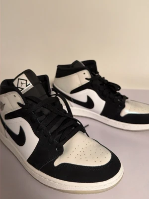 Nike Air Jordan 1 svart/vit sneakers - Nike Air Jordan 1 sneakers i svart och vit med klassisk swoosh på sidan. Skorna har snörning, rund tå och platt sula. Materialet är en mix av skinn och mocka, med svart ovandel och vita paneler. Perfekt för dig som gillar ikoniska och stilrena sneakers.