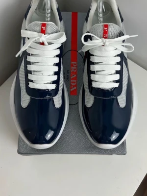 Prada sneakers i marinblå och vit - Säljer ett par snygga Prada sneakers med marinblå lackdetaljer och vit mesh. Skorna har vit snörning, rund tå och en platt sula. Röd logga på plösen ger en exklusiv touch. Perfekta för dig som gillar stilrena och lyxiga sneakers.