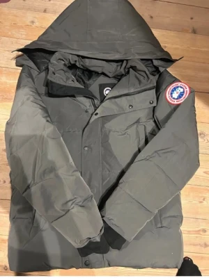Mörkgrå dunjacka från Canada Goose - Säljer en mörkgrå dunjacka från Canada Goose med klassisk patch på ärmen. Jackan har huva, långa ärmar och stängs med både dragkedja och knappar. Perfekt för kalla dagar och riktigt snygg streetstil. Använd några dagar bara!. Kontakta mig för en video när jag har på mig den!