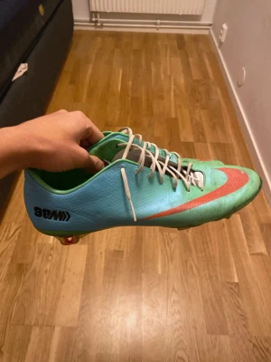 Nike fotbollsskor turkos och orange - Säljer ett par Nike fotbollsskor i turkos och orange med vit snörning och låg profil. Skorna har en rund tå och platt sula med dobbar för gräsplan. Snyggt Nike-märke på sidan och hälen, tillverkade i syntetmaterial för lätt känsla på foten.