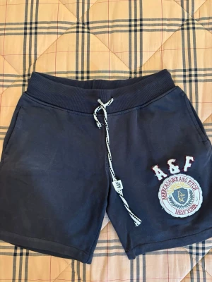 Mörkblå shorts från Abercrombie & Fitch - Säljer ett par mörkblå shorts från Abercrombie & Fitch med broderad logga och emblem på ena benet. De har snörning i midjan och är gjorda i mjuk bomull. Perfekta för chill dagar och har en loose passform.