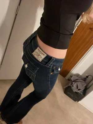 True Religion mörkblå bootcut jeans Joey  - Säljer ett par mörkblå jeans från True Religion med bootcut-snitt och låg midja. Jeansen har snygga detaljer som stora fickor med knappar och kontrastsömmar. Materialet är klassisk jeans och passformen är normal. Perfekta för dig som gillar en cool och avslappnad stil. Skriv för fler bilder. Dem är även uppstöttade längst ned, pris går även att diskutera 🥰