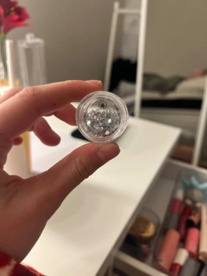 Silverfärgade ansiktsstenar Shine Stories - Säljer ett litet plastburk med silverfärgade ansiktsstenar från Shine Stories. Perfekt för att skapa kreativa och glittriga sminklooks. Stenarna är i olika storlekar och kan användas runt ögonen eller i ansiktet för extra effekt.