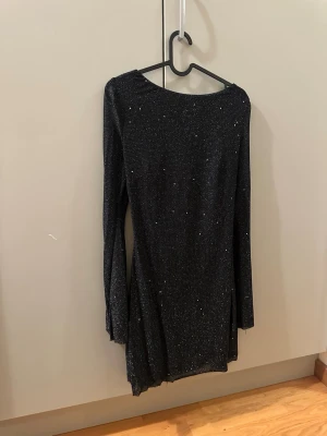 Svart glittrig klänning med öppen rygg - Svart glittrig kortklänning med långa ärmar och en djup öppen rygg som knyts med ett band. Klänningen har ett glittrigt mönster och är figurnära med lätt stretchigt material. Perfekt för dig som vill sticka ut med en snygg och trendig look. Pass utmärkt för festligheter! Endast använd en gång!