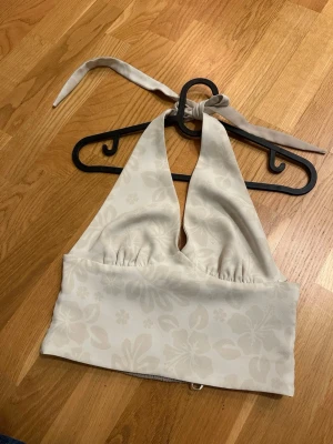Beige topp - Säljer en ljusbeige halterneck topp med subtilt blommönster. Toppen har bred smockad kant nedtill i bak och knytband i nacken. Materialet känns mjukt och svalt, perfekt för varma dagar. Passformen är croppad och toppen är ärmlös.