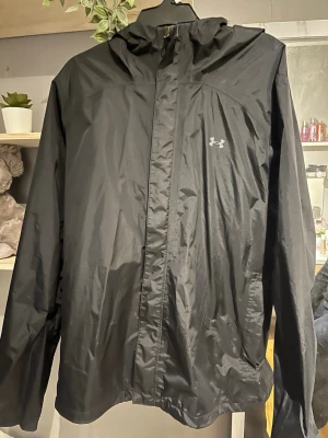 Svart vindjacka från Under Armour - Snygg svart vindjacka från Under Armour med huva och dragkedja framtill. Jackan har en minimalistisk look med diskret logga på bröstet och är tillverkad i lätt syntetmaterial. Perfekt för blåsiga dagar och enkel att slänga på sig.