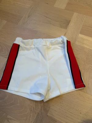 Vita sportiga shorts med röd sidrand - Snygga vita shorts med elastisk midja och röda samt svarta ränder längs sidorna. Perfekta för dig som gillar en sportig look och vill ha något bekvämt och stilrent. 