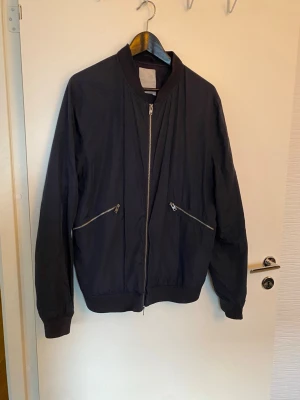 Mörkblå bomberjacka från J.Lindeberg - Mörkblå tunn bomberjacka från J.Lindeberg med snygga silverfärgade dragkedjor på fickorna och framtill. Jackan har ribbade muddar vid ärmslut och midja, samt är tillverkad i 100% polyester. Perfekt för dig som gillar stilren och enkel design.