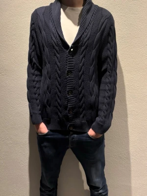 Mörkblå stickad kofta Hampton Republic - Säljer en mörkblå, grovstickad kofta från Hampton Republic i storlek M. Koftan har snygga kabelstickade detaljer, bred krage och knäppning med fyra knappar framtill. Perfekt för lager-på-lager och chill stil under höst och vinter. Knappen längst upps hål, är lite slitet (syns på bild), därför ibland släpps den av, men inte jätte ofta. Storlek M (medium), men skulle säga den passar bättre en L (large)