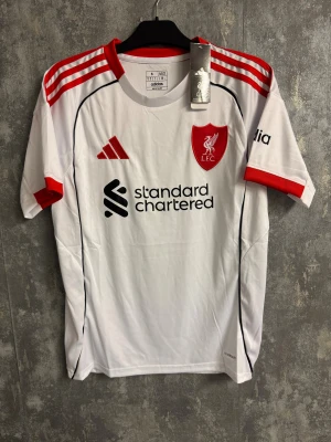 Vit liverpool tröja - Ny storlek S - Bilderna visar en vit och röd Liverpool FC fotbollströja från Adidas, inte skor. Tröjan har röda detaljer och klubbmärke på bröstet.