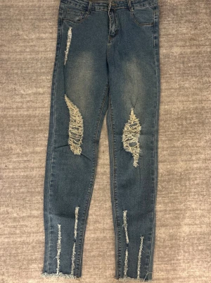 Blå ripped jeans - Säljer ett par blå jeans med slitna detaljer och stora ripped hål på benen. Jeansen har fransade benslut och klassisk femficksdesign. Modellen är smal och sitter tajt längs benen.