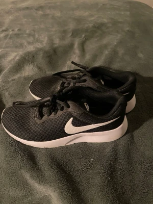 Svarta Nike sneakers strl 38 - Säljer ett par svarta Nike sneakers med vit sula och vit swoosh på sidan. Skorna har mesh-överdel för bra ventilation och snörning framtill. Snygg och sportig design som passar perfekt till streetwear och träning. Sulan är platt och har tydligt Nike-märke undertill.