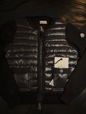 Moncler Cardigan  - Snygg svart pufferjacka från Moncler med quiltad framsida och släta ärmar. Jackan har dragkedja, två fickor med knappstängning och Moncler-logga på ärmen. Perfekt för dig som vill ha en stilren och modern look.