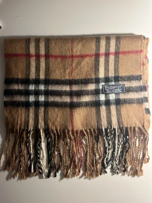 Burberrys rutig kashmirväst - Säljer en klassisk rutig väst från Burberrys i 100% kashmir. Västen har beige, svart, vit och röd färg med det ikoniska rutmönstret och fransar längst ner. Perfekt för dig som gillar tidlös design och mjuka material. (10C)