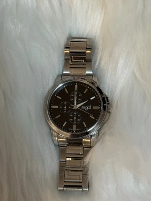 Regel Ur&Penn Klocka  - Säljer en stilren silvrig klocka från Regal med svart urtavla och metallarmband, inga fel men behöver byta ut batteri. 