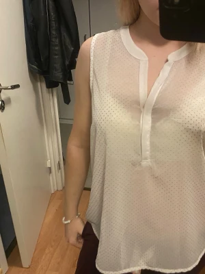 Vit prickig ärmlös blus - Säljer en luftig och transparent vit blus med små diskreta prickar. Blusen är ärmlös och har en snygg v-ringad halsringning med bred kant. Perfekt till sommaren och enkel att styla med olika accessoarer.