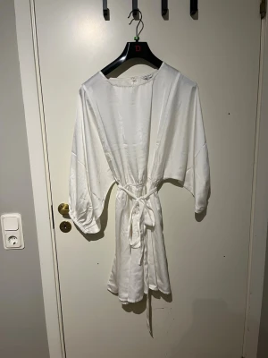 Vit kortklänning  - Säljer en vit kortklänning med rund hals, trekvartsärmar och knytband i midjan. Klänningen har en mjuk, glansig yta som ger en lyxig känsla och är tillverkad i ett silkesliknande material. Perfekt för dig som gillar stilrena och eleganta plagg. Använd en gång 