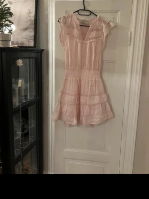 Ljusrosa kortklänning från Neo Noir - Supersöt ljusrosa klänning från Neo Noir med volanger och spetsdetaljer. Modellen är kort med v-ringad hals och ärmlös design. Tillverkad i 100% bomull med mjuk viskosfoder. Perfekt för sommarens alla tillfällen!