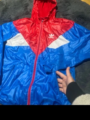 Adidas färgglad vindjacka med huva - Säljer en snygg Adidas vindjacka i rött, blått och vitt med coolt geometriskt mönster. Jackan har huva med snörning, dragkedja framtill och fickor med dragkedja. Materialet är lätt och syntetiskt, perfekt för blåsiga dagar. Adidas-logga på bröstet.