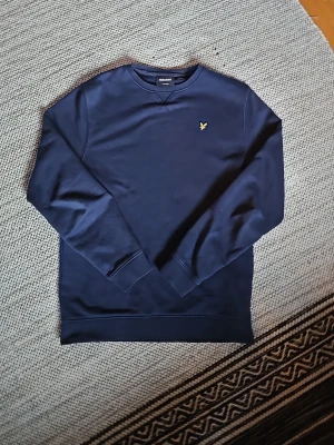 Marinblå sweatshirt från Lyle & Scott - Säljer en stilren marinblå sweatshirt från Lyle & Scott med klassisk rund hals och gul broderad logga på bröstet. Tröjan har långa ärmar och ribbade muddar vid ärmslut och nederkant. Perfekt för dig som gillar enkel och snygg design.