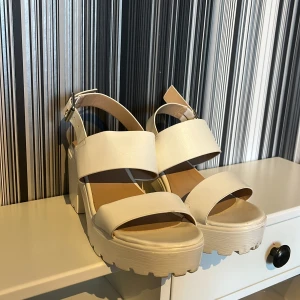Vita chunky sandaler från Tiamo - Säljer ett par vita sandaler från Tiamo med breda remmar och justerbart spänne vid ankeln. Använda en gång vid en skolavslutning. Sandalerna har en chunky plattformssula med grovt mönster undertill. Materialet är syntet och de har öppen tå.