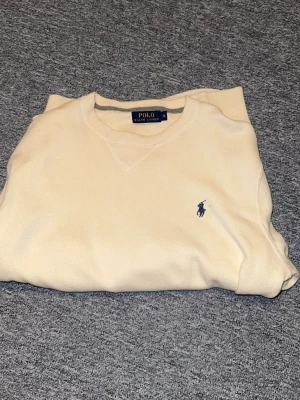 Beige stickad tröja Polo Ralph Lauren - Snygg beige stickad tröja från Polo Ralph Lauren i storlek S. Klassisk rund hals och långärmad modell med den ikoniska blå Polo-loggan broderad på bröstet. Perfekt för dig som gillar stilrena och tidlösa plagg.