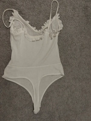 Vit ribbad bodysuit med volang - Säljer en vit ribbad bodysuit med smala axelband och volangdetaljer längs v-ringningen. Materialet är mjukt och stretchigt, perfekt för sommarens alla tillfällen. Bodyn är ärmlös och har en feminin touch med volangerna.