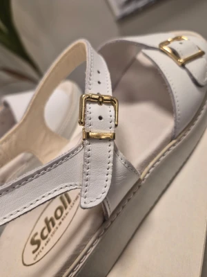 Nya! Scholl 651 vit läder med guldiga detaljer sandaler EUR 39 - Scholl sandaler i vitt skinn med guldfärgade spännen och öppen tå. Snygg platt sula och klassisk design med bred rem över foten och justerbar rem runt ankeln. Perfekta för dig som gillar stilrena och bekväma skor med lite retrovibe.