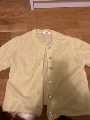 Ljusgul stickad kofta från Monki - Säljer en ljusgul stickad kofta från Monki med rund hals och silvriga knappar framtill. Koftan har lång ärm och är gjord i ett mjukt stickat material som känns skönt mot huden. Perfekt att slänga över en t-shirt när det är lite kyligt ute.