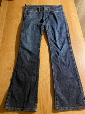 Blå bootcut jeans lågmidjade - Säljer ett par klassiska bootcut jeans i mörkblå denim med låg midja. Byxorna har fem fickor, bälteshällor och snygga kontrastsömmar. Märkespatch på bakfickan och slitstark bomullskvalitet. Perfekt för dig som gillar retrostil och bredare benslut.